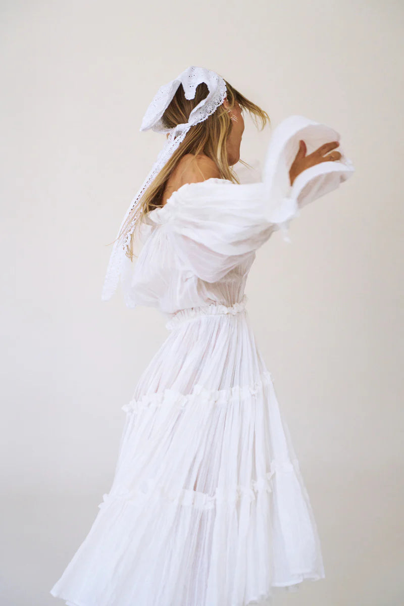A woman wearing a white off-the-shoulder tiered dress with a matching white bonnet and veil, photographed against a plain light beige background.