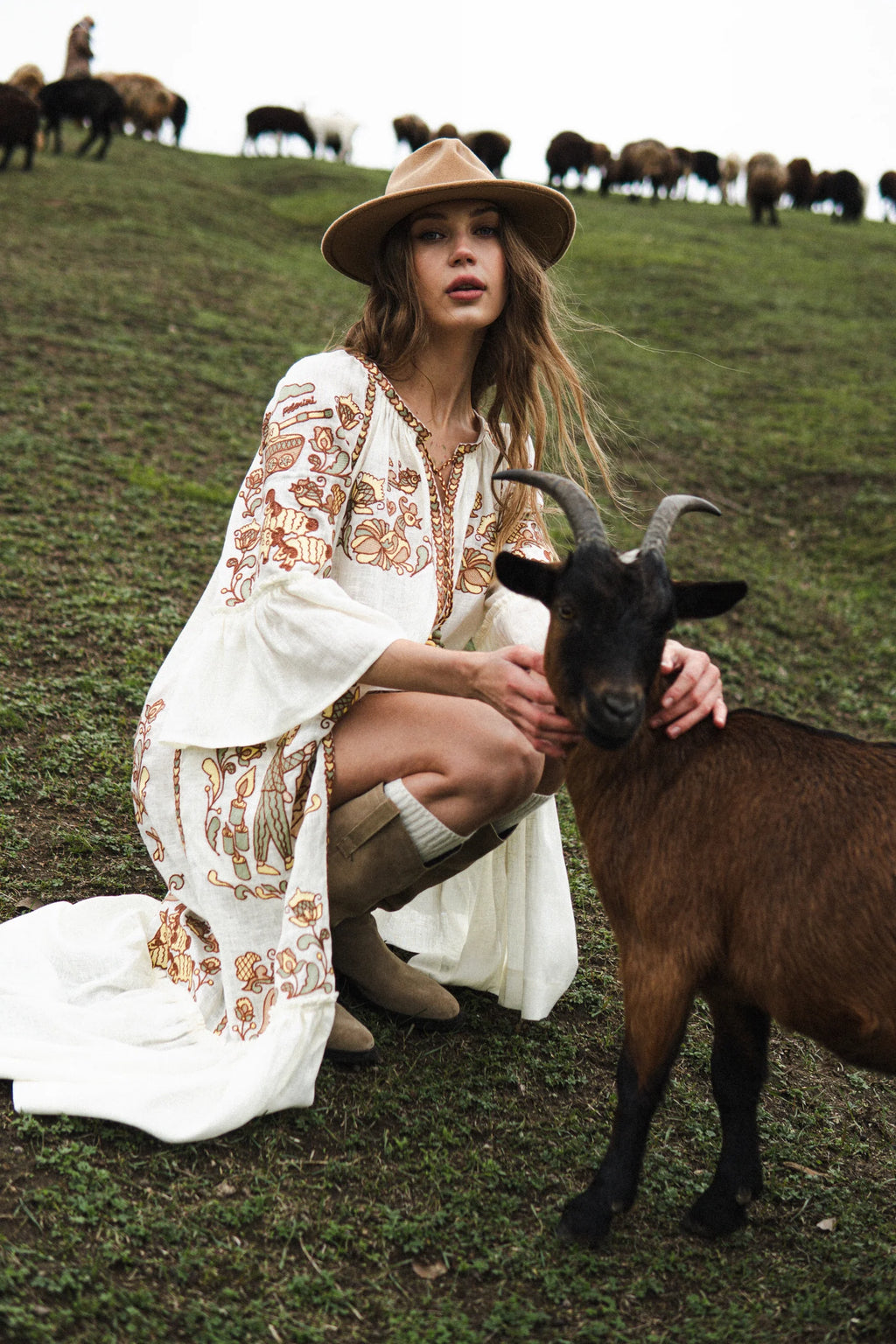 Woman in a white embroidered dress and hat with a goat on a grassy hill with grazing animals in the background