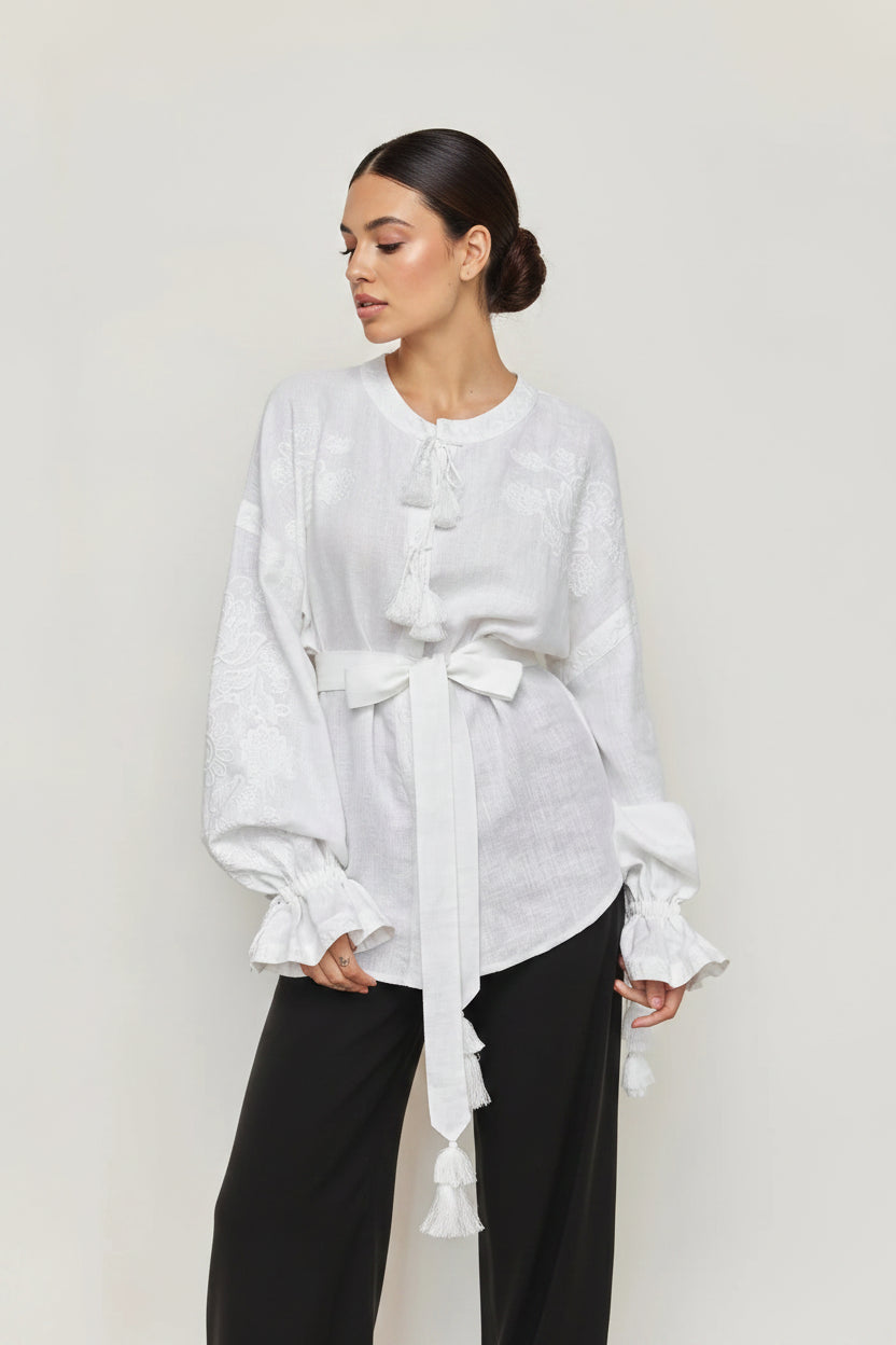 A woman modeling a white long-sleeve blouse with a tie waist, tassel details, and ruffled cuffs, paired with black pants, against a plain light gray background.