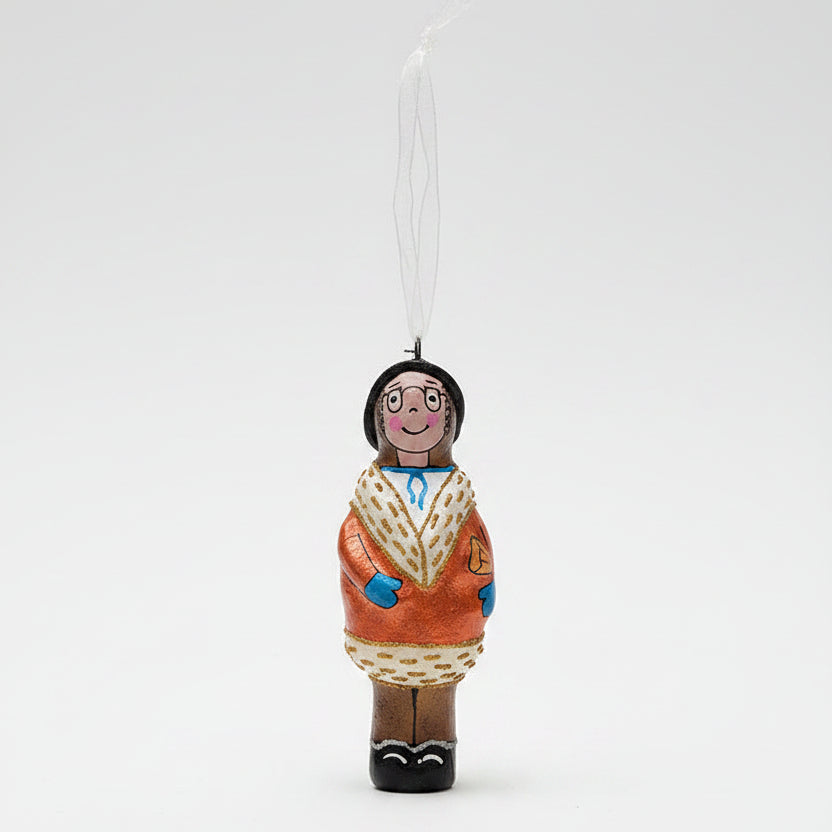 A hand-painted wooden Christmas ornament shaped like a person in a patterned coat, hanging from a pine branch against a dark green background and cream surface.