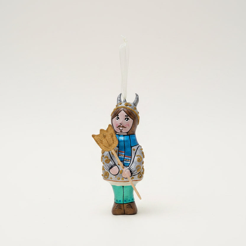 A hand-painted wooden Christmas ornament shaped like a knight in armor holding a sword, hanging from a pine branch against a dark green background and cream surface.