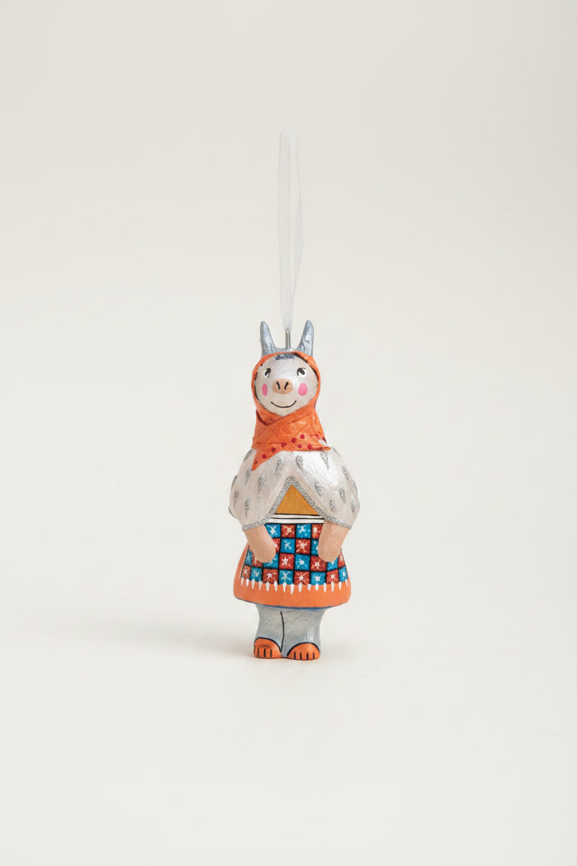 A hand-painted llama Christmas ornament hanging from a pine branch with a white ribbon, set against a rustic beige and brown background with draped white fabric.