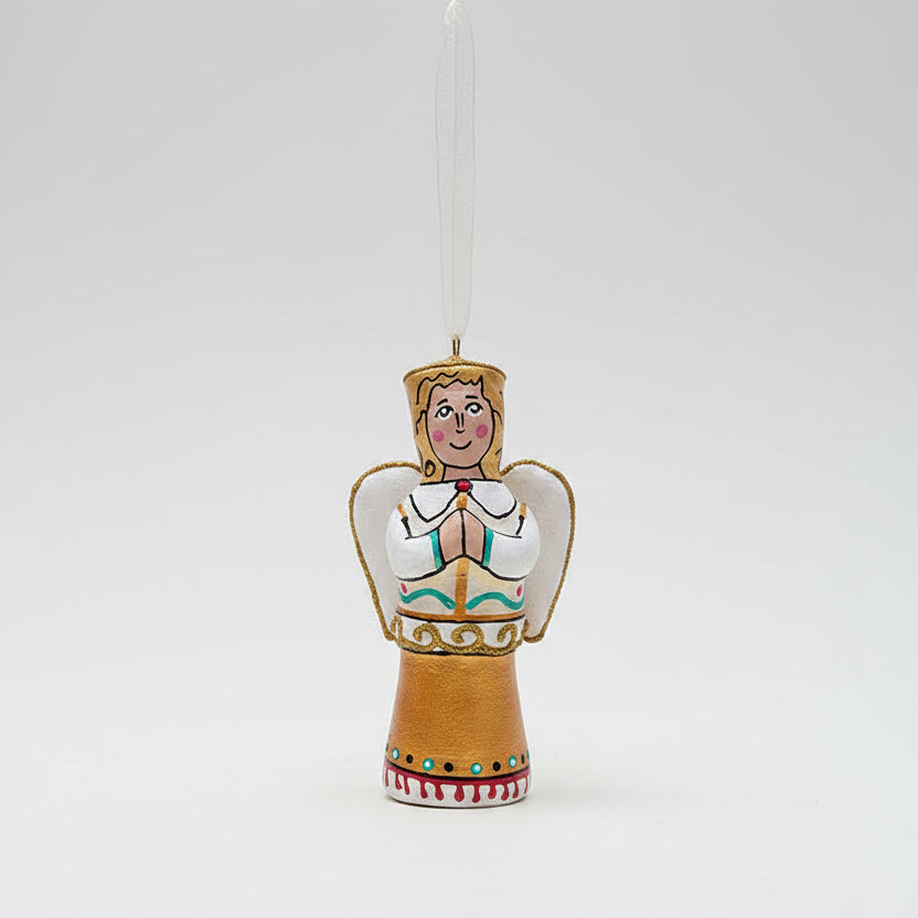 A hand-painted wooden Christmas angel ornament hanging from a pine branch against a dark green background and cream-colored surface.