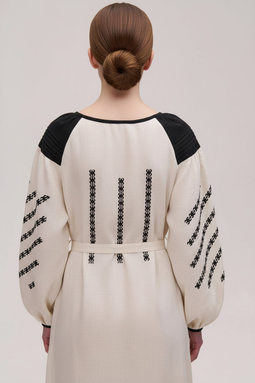 Person wearing a white dress with black embroidery on a plain background