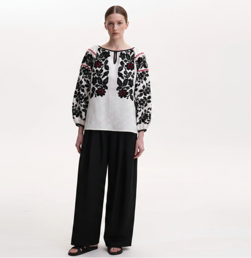 A model wearing a white long-sleeve top with black floral embroidery and red accents, paired with black wide-leg trousers, against a plain white studio background.